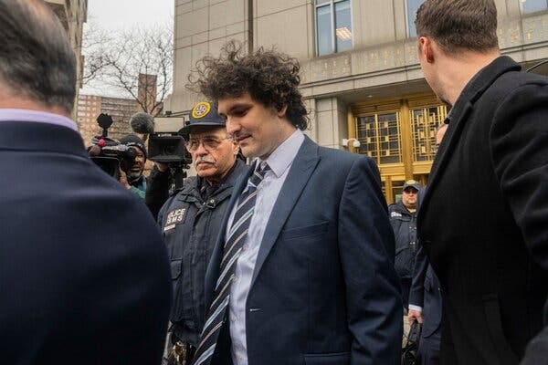 Sam Bankman-Fried walking away from a building with gold door and window frames, while surrounded by people and cameras.