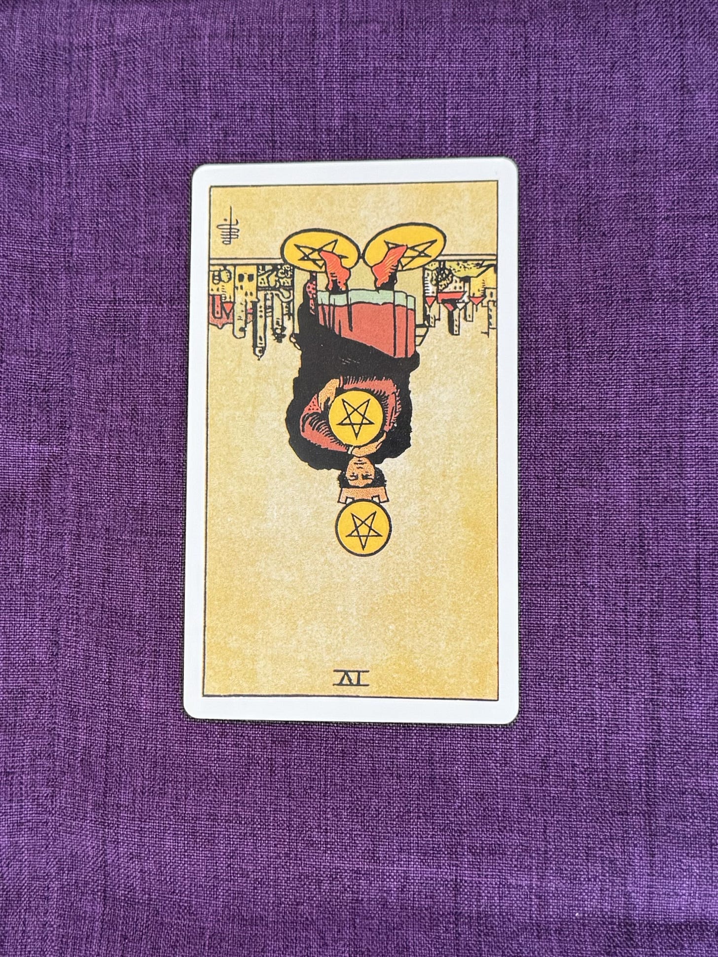A figure sits on a stone bench outside a walled city. A golden coin rests on top of the figure’s head, one coin is held against the chest with both arms, and one coin is positioned beneath each foot. The figure wears a crown and a long cloak. Buildings with towers are visible in the background beneath a clear sky. The card is upside down on a purple linen background.