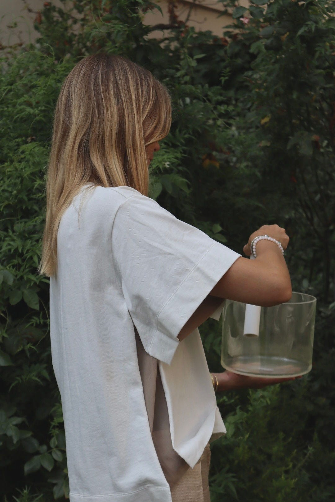 Woman seen from the side with long blonde hair and a white oversized T-shirt, holding a clear crystal singing bowl and a mallet in a green garden. Woman seen from the side with long blonde hair and a white oversized T-shirt, holding a clear crystal singing bowl and a mallet in a green garden.
