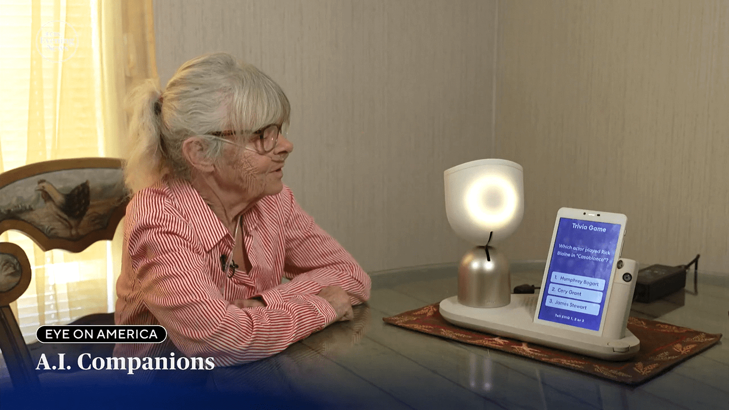 An older woman sits at a table facing the ElliQ robot with an attached tablet showing a trivia game.