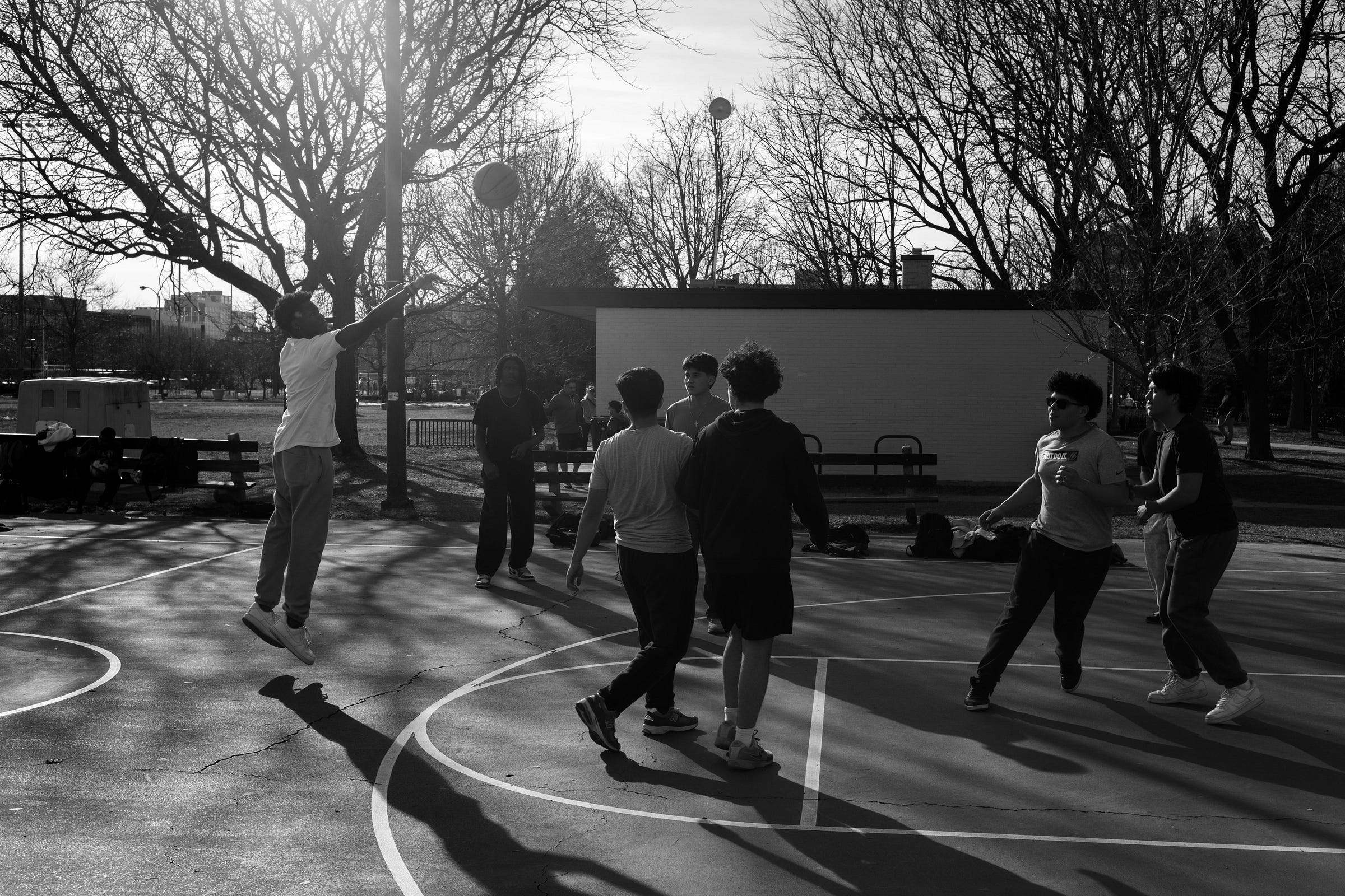 A black-and-white photograph of a group of young men playing basketball on an outdoor court in the late afternoon. One player is mid-air, releasing a jump shot, his shadow stretching across the court. Others watch, poised to react, while sunlight filters through the leafless trees, casting long shadows and highlighting the movement and energy of the game. In the background, a small brick building, park benches, and a cityscape set the scene.