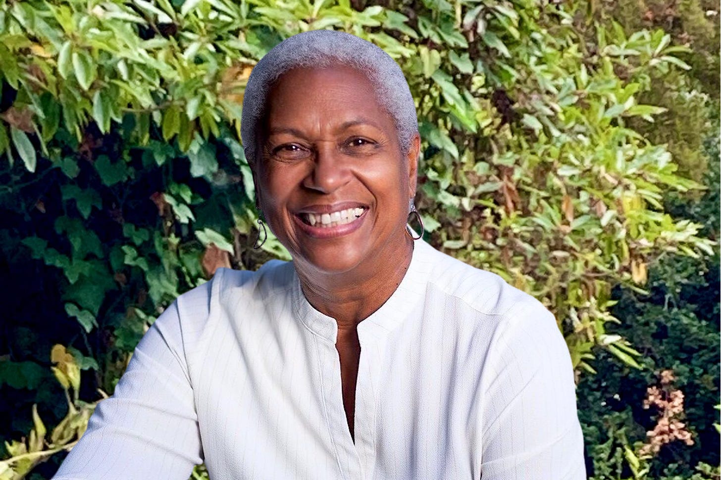 Akaya, an African American woman with short white hair, smiles at the camera. Behind her are the green leaves of trees and bushes.