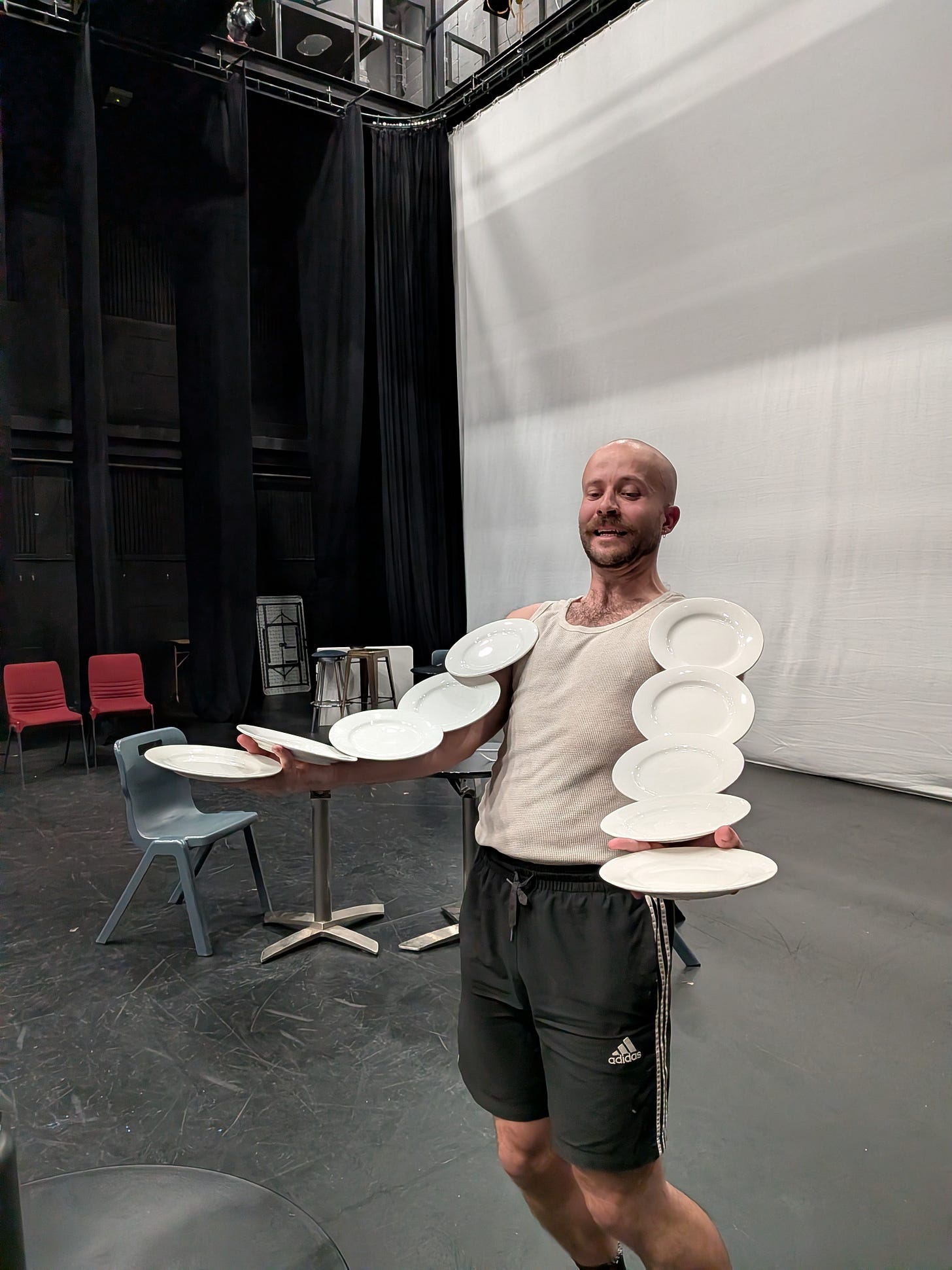 Lachlan standing with arms extended, balancing multiple white plates on each arm in the Bruce Gordon theatre. Behind Lachlan are black stage curtains, chairs, and tables.