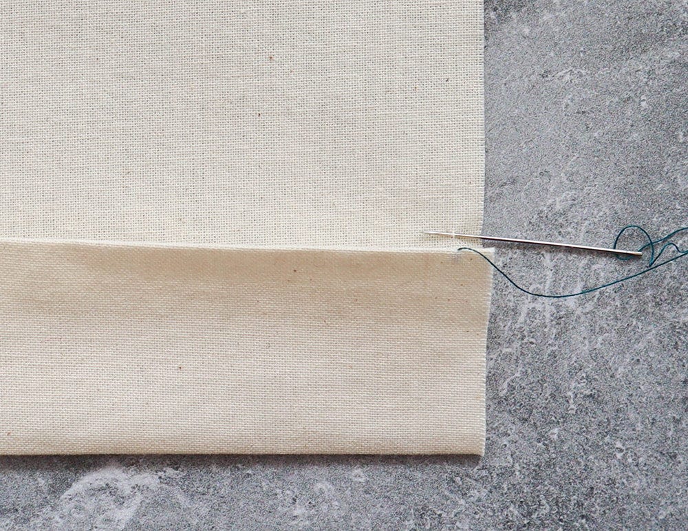 Cotton fabric with hem folded up and needle with teal thread on concrete surface