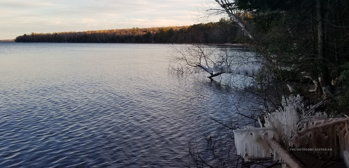 A landscape photograph taken on a late winter or early spring evening at the edge of Frog Bay. The open, calm water reflects the twilight sky, looking toward the distant horizon where the tip of Hermit Island sits. In the immediate foreground, thick icicles hang gracefully from low-hanging tree branches over the sandy beach.