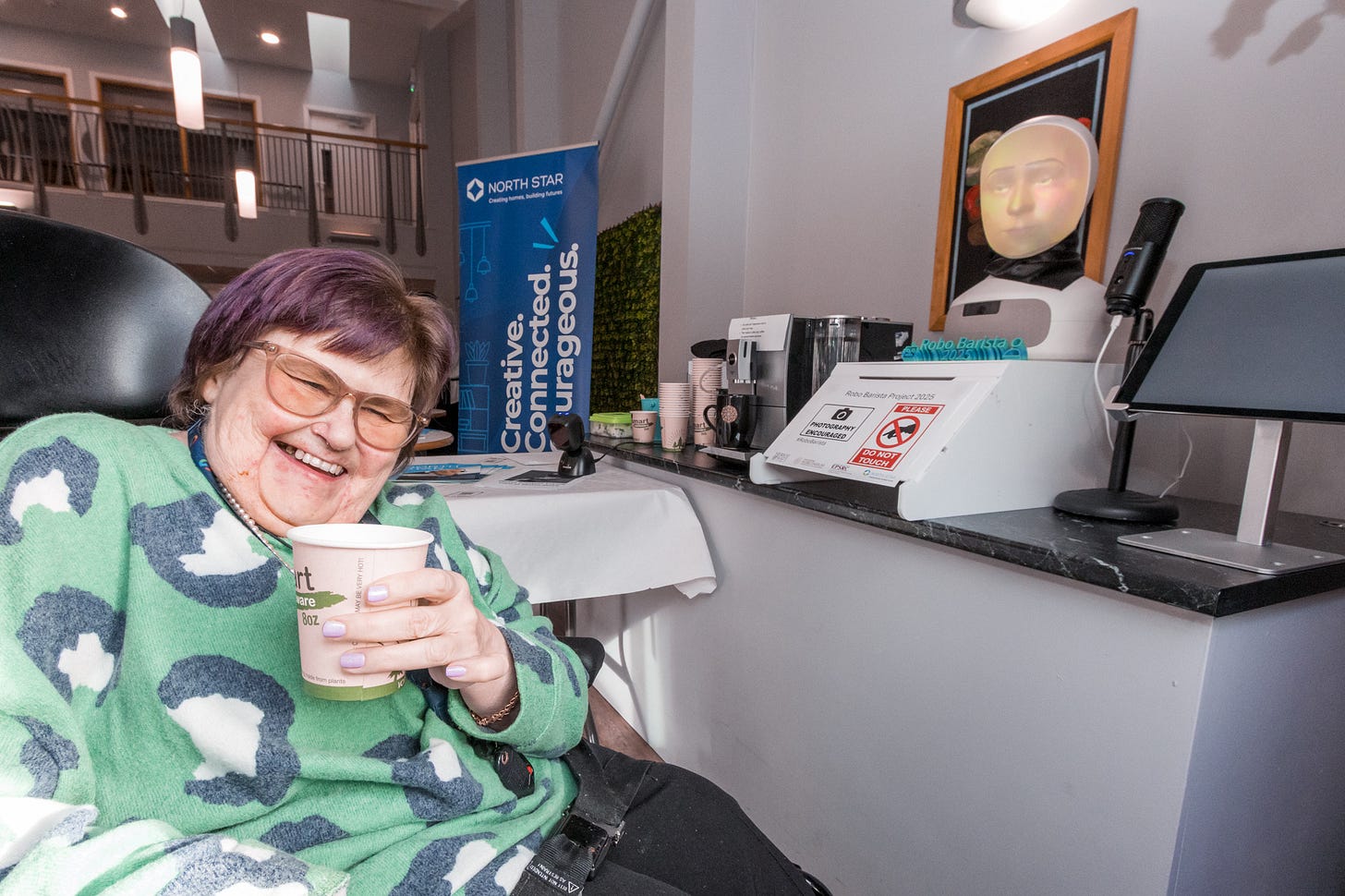 An older woman wearing a green jumper with a very large leopard print pattern on it sits in a chair holding a coffee cup. Next to her, glowing brightly, is a robotic head with a face projected on the inside. In the background a banner reads "North Star Housing Group. Creative. Connected. Courageous."