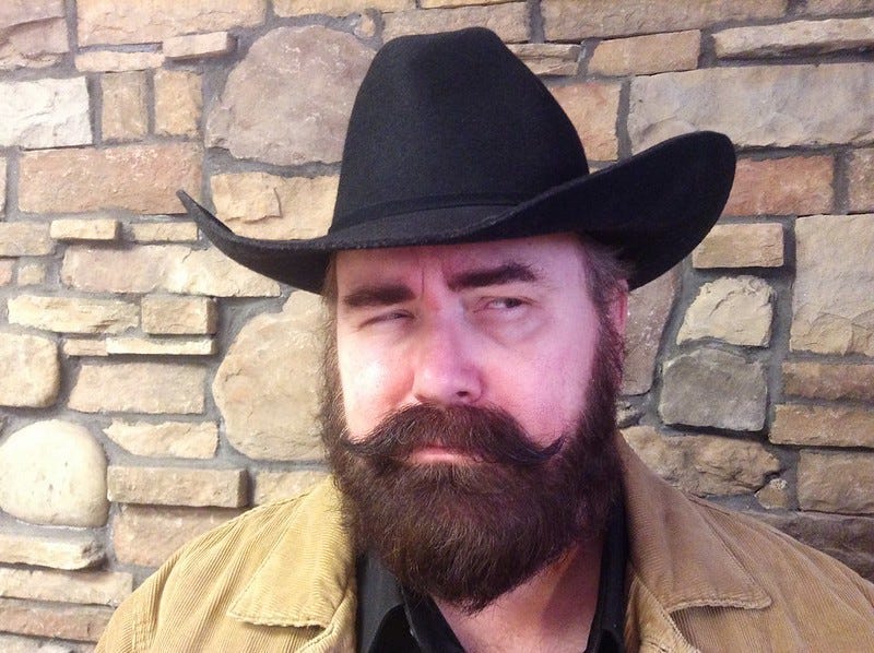 A bearded man with a mustache and cowboy hat against a stone wall squinting in suspicion.