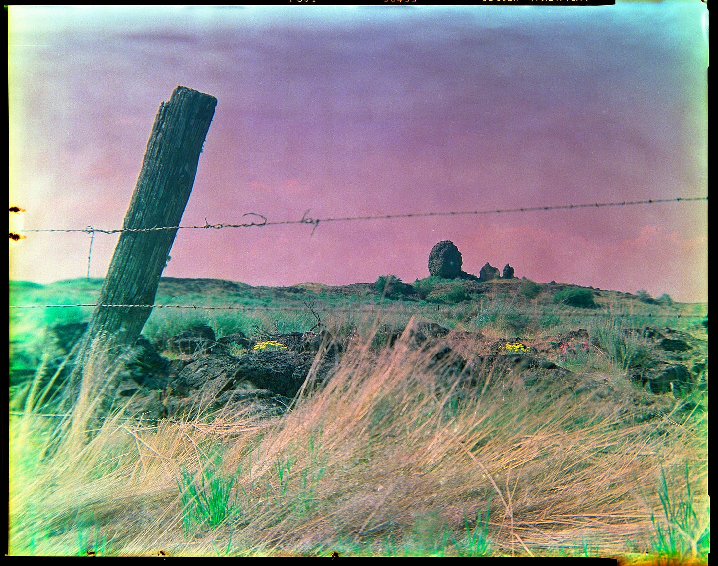 A large basalt boulder rests on the horizon. The photo is taken looking through a barbed wire fence. 