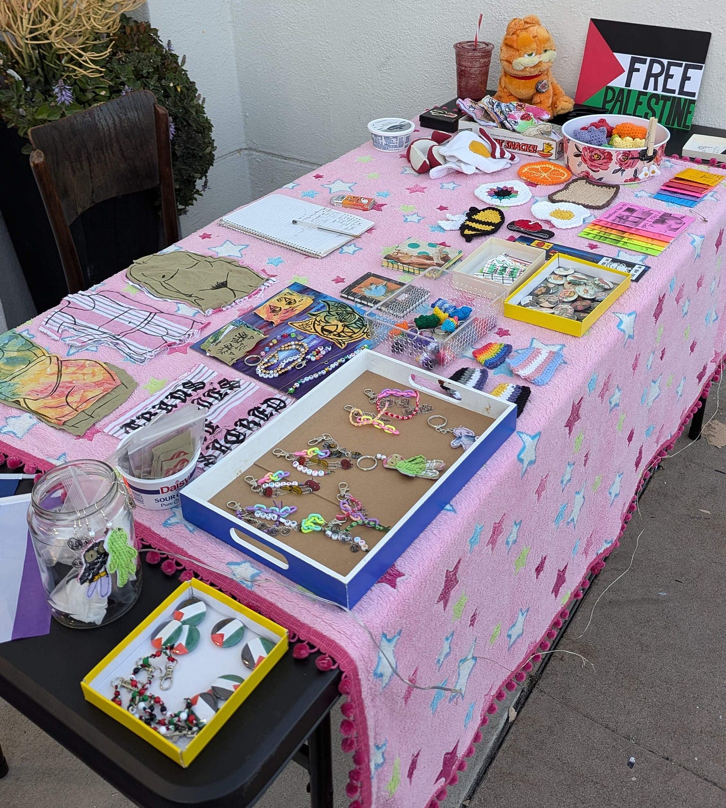 A table with a wide variety or art object laid out on it. There are keychains, zines, patches, stickers, and buttons. All are colorful and many are related to trans or queer pride. There is also a "Free Palestine" sign on the table.