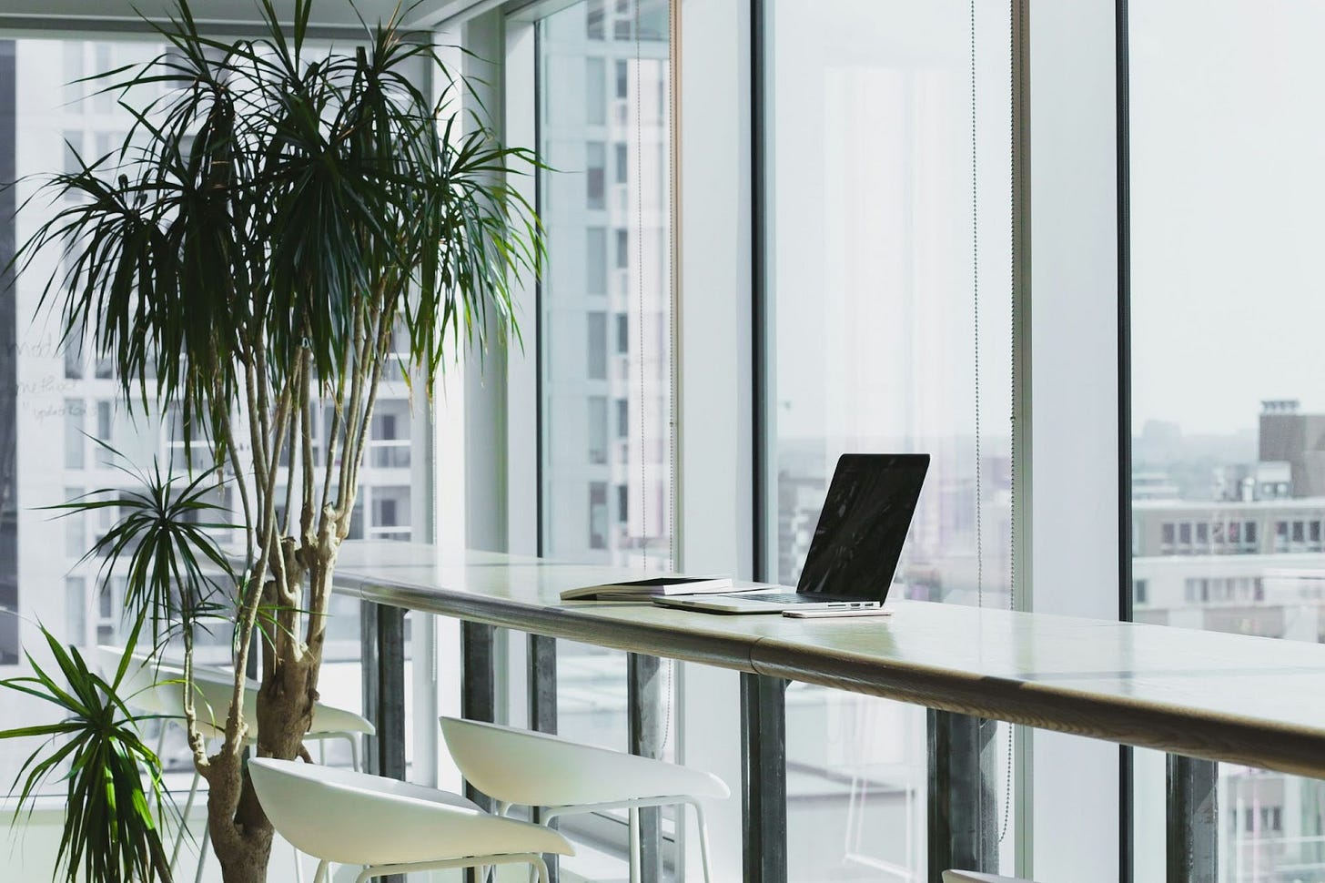 Laptop on a table along the glass windows of a high-rise office with a plant in the corner. Laptop on a table along the glass windows of a high-rise office with a plant in the corner.