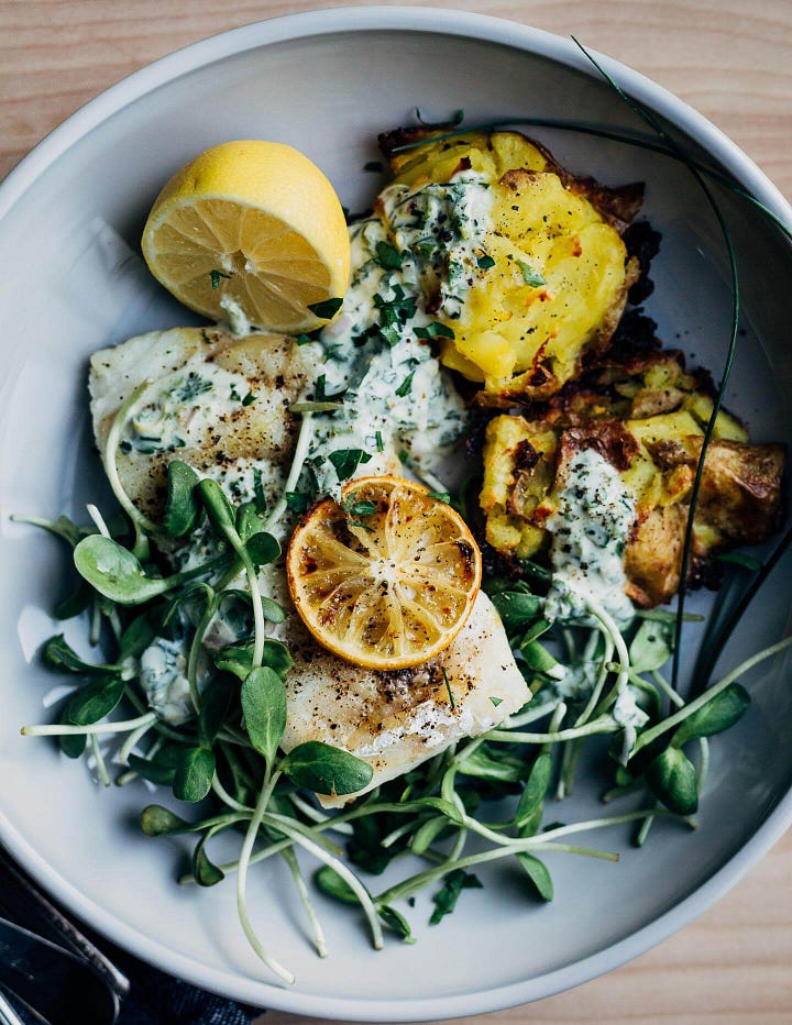 On the left, a wide bowl with smashed potatoes, cod, roasted lemons, microgreens, and a creamy sauce. On the right, a skillet with hands tossing noodles with shrimp and pesto.