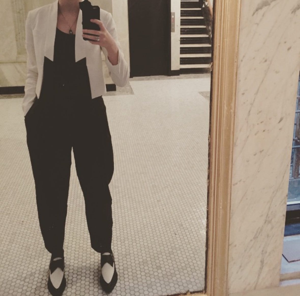 woman, shown from neck to feet, taking a selfie. she is in a black jumpsuit with a white blazer. woman, shown from neck to feet, taking a selfie. she is in a black jumpsuit with a white blazer.