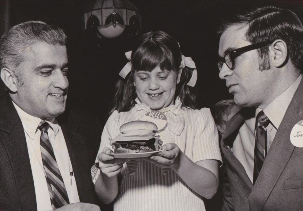 A vintage black-and-white photo of a young girl smiling and holding a hamburger on a tray, flanked by two suited men. The man on the right wears glasses and a name tag reading “John.” This image is from the early days of Wendy’s. A vintage black-and-white photo of a young girl smiling and holding a hamburger on a tray, flanked by two suited men. The man on the right wears glasses and a name tag reading “John.” This image is from the early days of Wendy’s.
