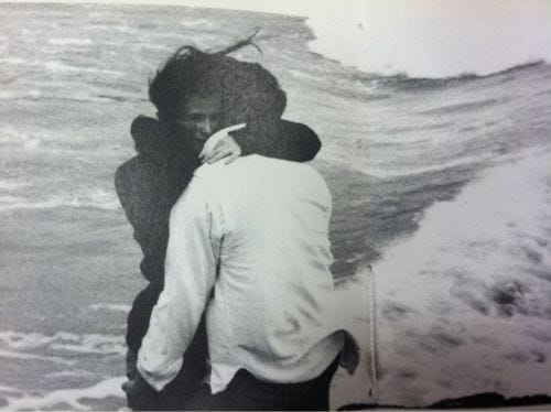 This may contain: two people hugging each other on the beach with waves crashing in front of them and one person holding his head