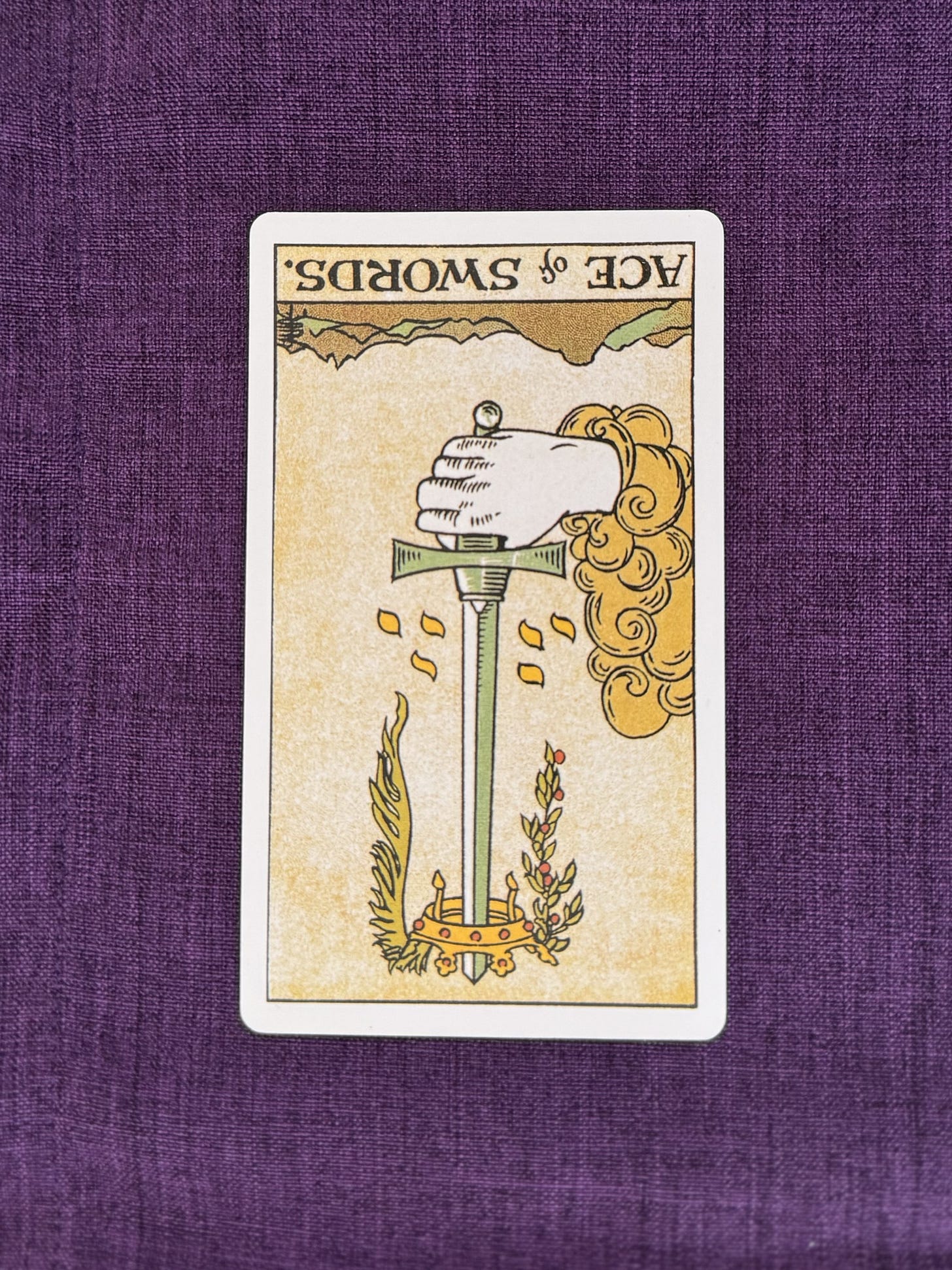 A hand emerges from a cloud, holding a single upright sword by its hilt. A crown encircled by a laurel wreath and a palm branch rests near the tip of the sword, with small leaf-like shapes falling around it. Below, jagged mountains rise from the landscape. The sky is pale, and no people are present.