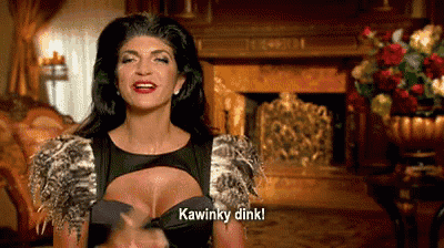 a woman in a black dress is standing in front of a fireplace and talking .