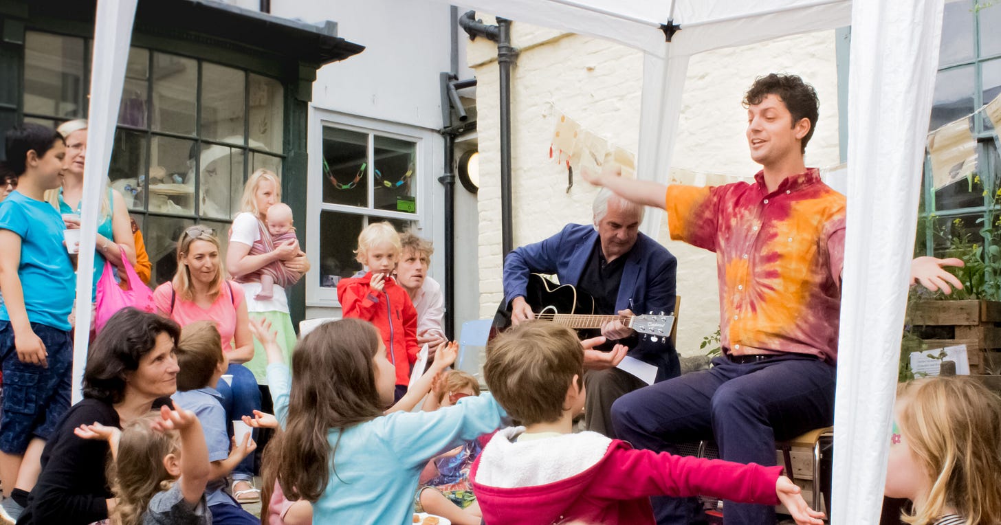 Storytelling to an all-age crowd in the courtyard of the Museum of Cambridge. I'm wearing a tie-dye shirt and sat slightly in front of a guitarist providing musical accompaniment. Many of the children have spread their arms own in imitation of my own gesture, showing their participation in my story.