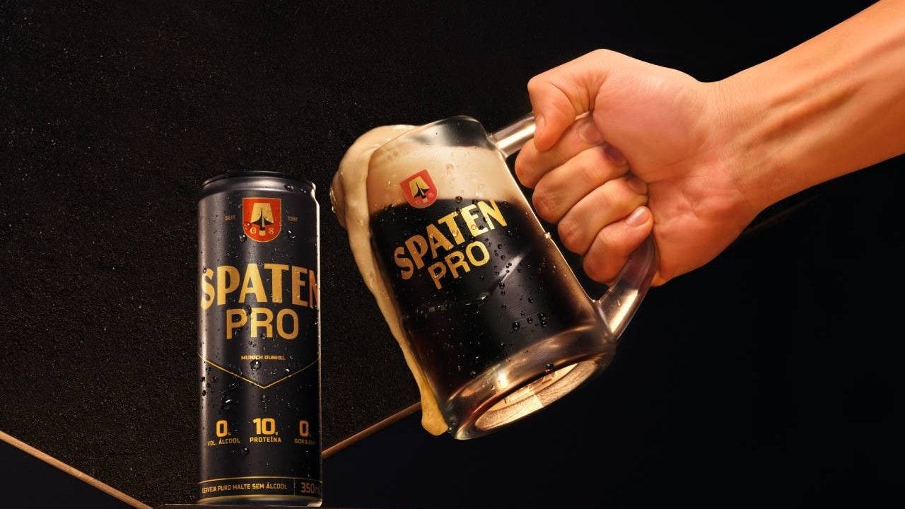 Uma lata preta e dourada de Spaten Pro e uma caneca de vidro cheia de cerveja escura com espuma transbordando, segurada por uma mão, em fundo preto. A lata exibe 0% vol. álcool, 10g proteína e 0g gordura.