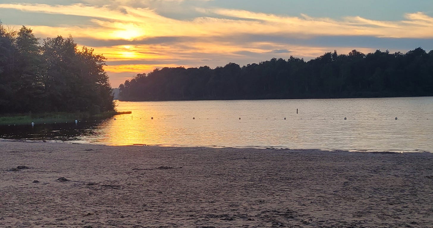 A sandy lake beach. The sun is setting behind clouds and trees.