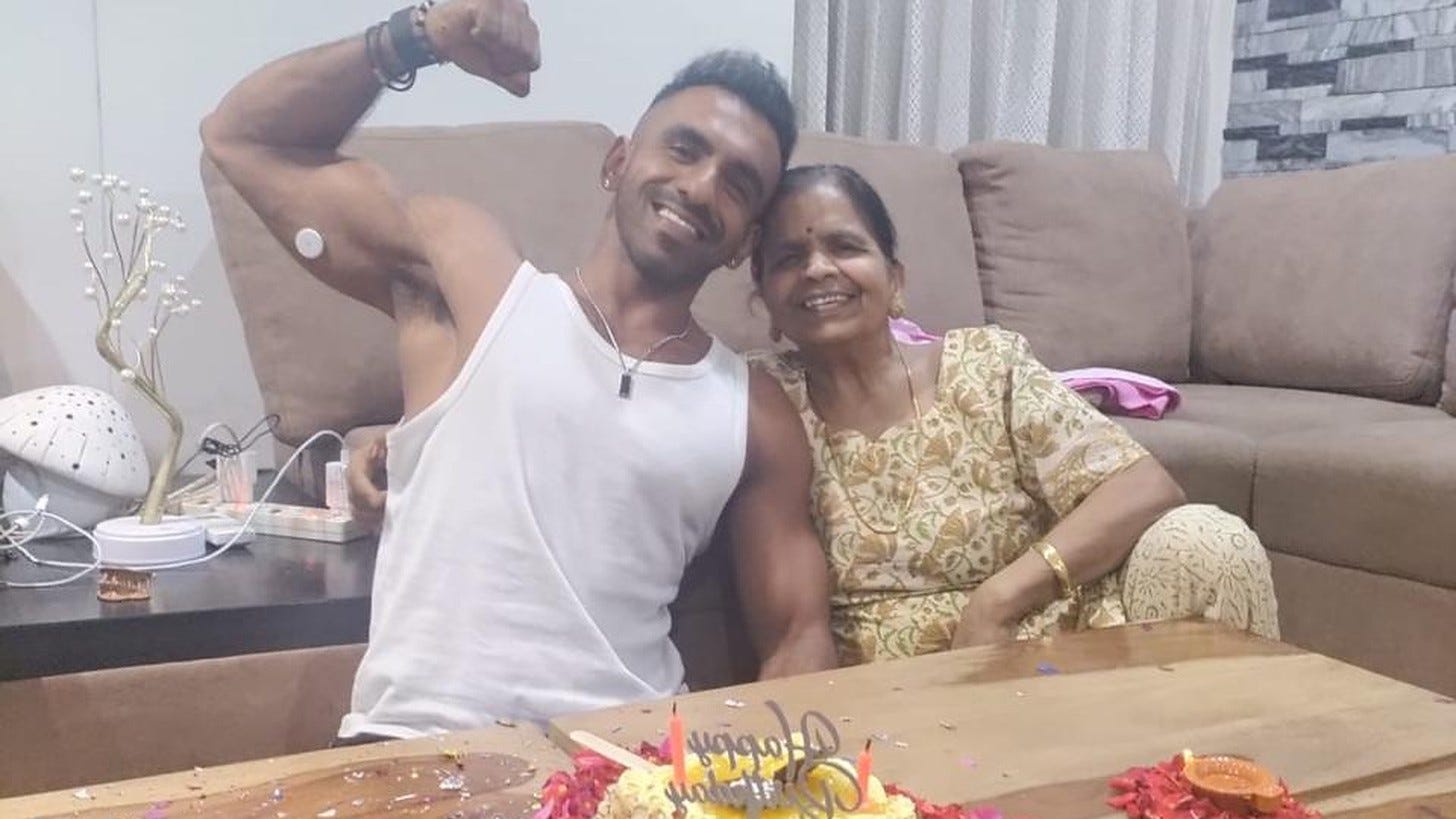 Avi flexing his arm and smiling beside an older woman on a sofa, a birthday cake with candles on the table in front of them. Avi flexing his arm and smiling beside an older woman on a sofa, a birthday cake with candles on the table in front of them.