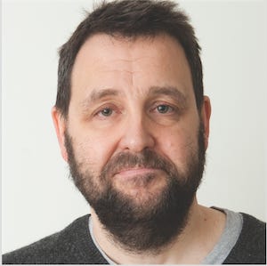 Image caption: A headshot of Russell. He’s a white man with short brown hair and a brown beard, wearing a grey jumper.
