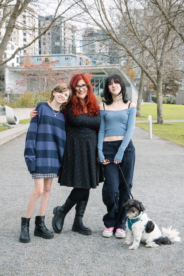 A redheaded mom and her two teen daughters stand in front of a modern building, outside, with their black and white dog. A redheaded mom and her two teen daughters stand in front of a modern building, outside, with their black and white dog.