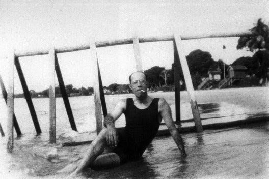 Fotografia preto e branco do escritor Mário de Andrade, meio careca, de óculos, e com um cigarro no canto da boca, de traje de banho preto, sentado bem despojado na beira da praia, posando na frente de uma estrutura de madeira. Ao fundo, a orla com casas e muitas árvores.