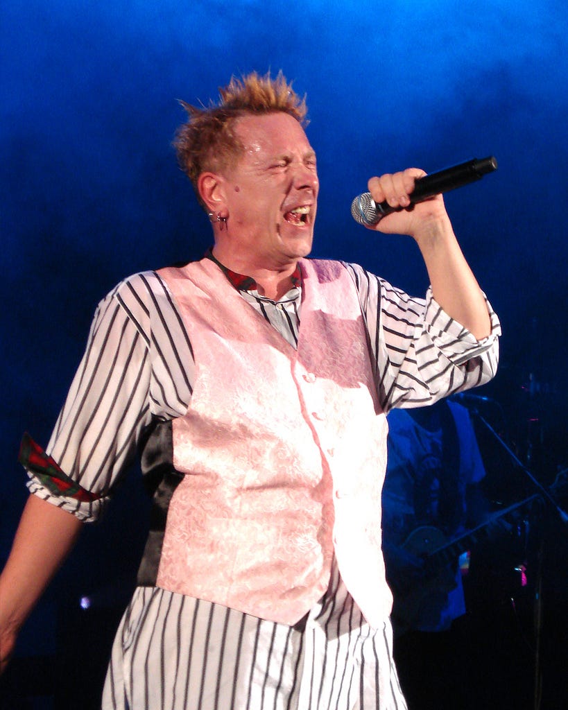 Lydon singing into microphone; spikey hair, pink vest, striped pants and shirt, face crinkled up Lydon singing into microphone; spikey hair, pink vest, striped pants and shirt, face crinkled up