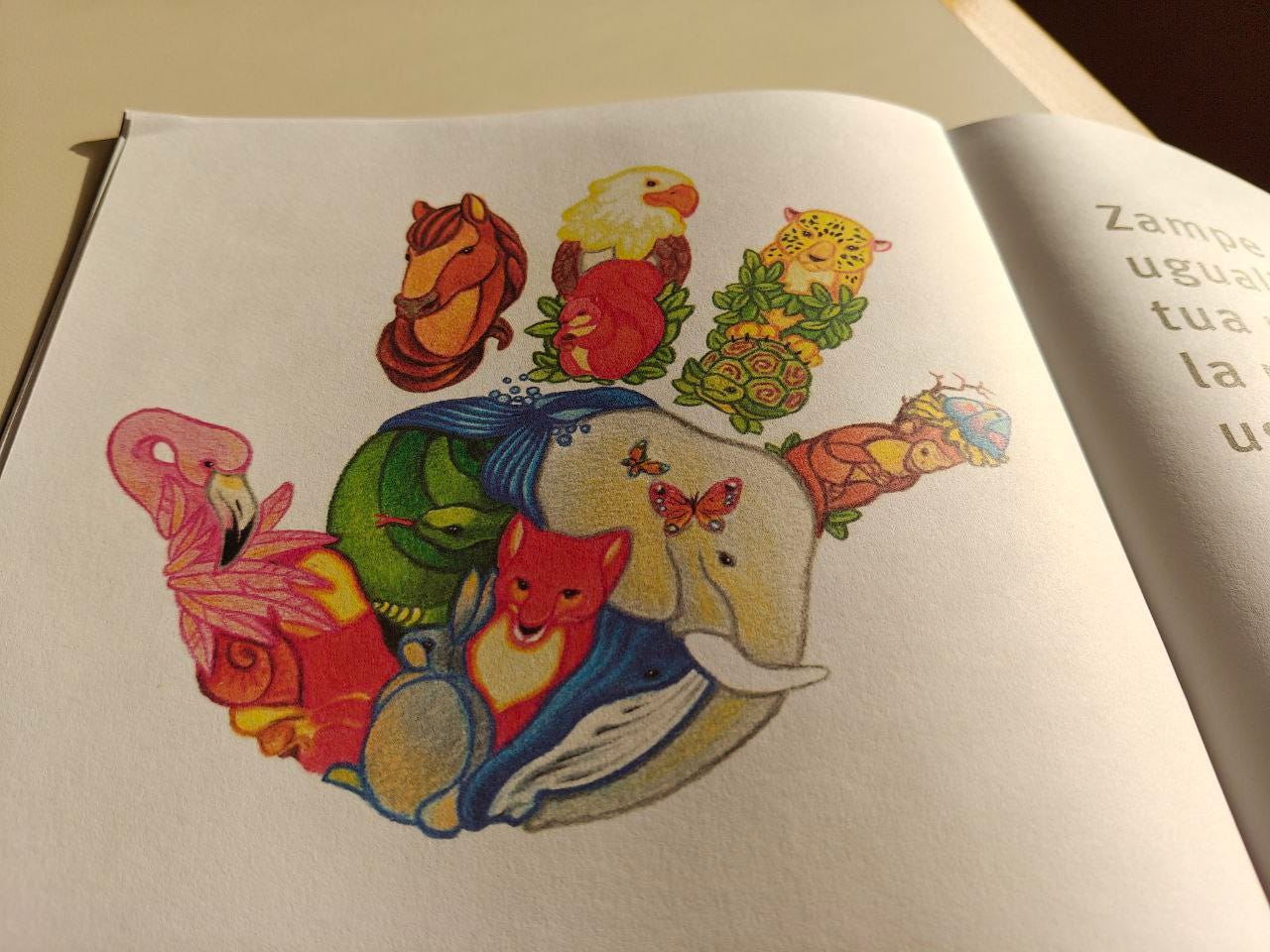 Una fotografia a colori mostra l'immagine di una mano aperta, dove ogni parte è composta da diversi animali vividamente illustrati e intrecciati tra loro, come in un mosaico. Tra gli animali visibili ci sono un fenicottero rosa (sul pollice), un cavallo (sull'indice), un'aquila o una calva (sul medio), un giaguaro o un leopardo (sull'anulare), e una serie di animali più piccoli sul palmo e sul polso, tra cui un elefante, una volpe, un serpente, una balena e due tartarughe. L'immagine è stampata su una pagina di un libro o quaderno, e sul lato destro si intravedono alcune parole in italiano.