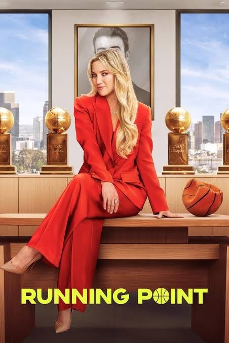 Kate Hudson sitting on a desk in a red suit with a basketball next to her