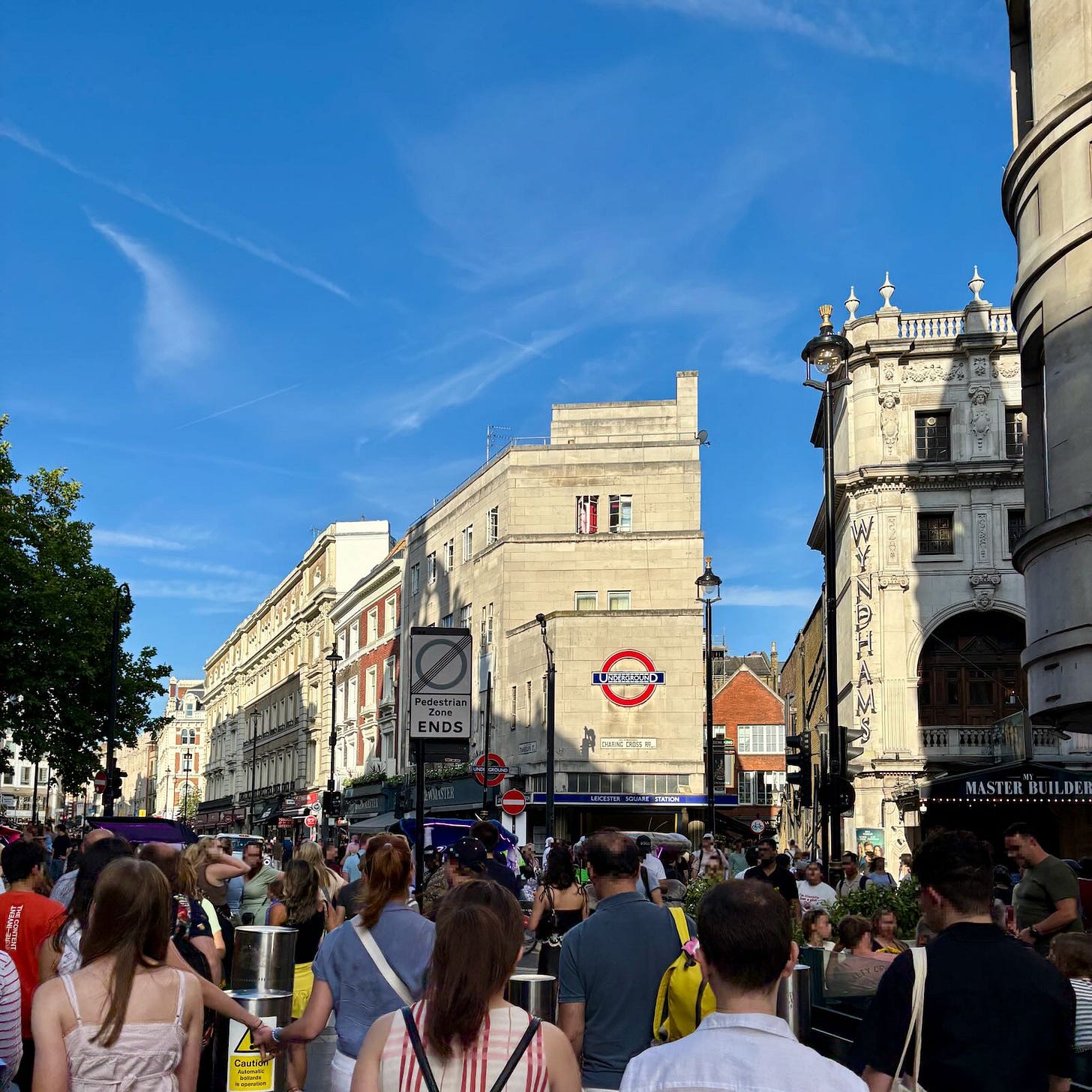 Crowd of people on a sunny evening on Cranbourne Street, London, with Leicester Square station visible in the distance.