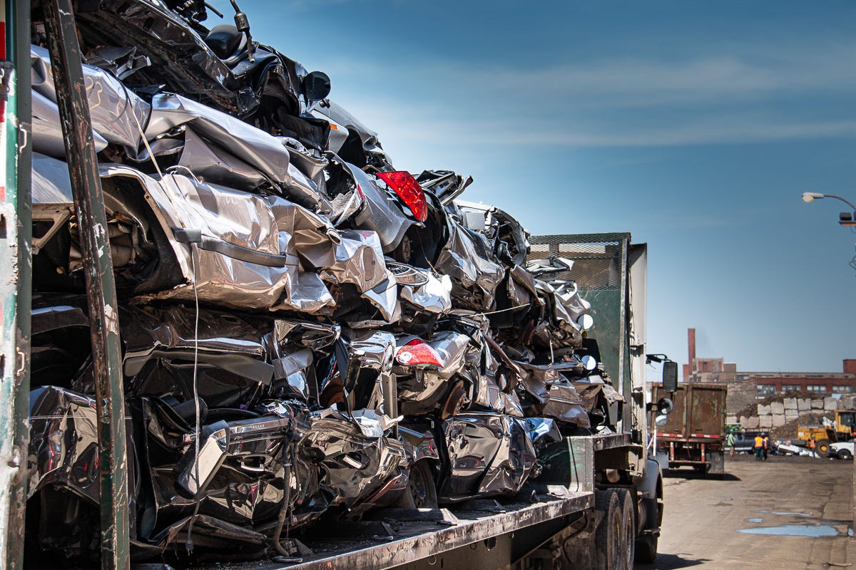 A trailer stacked with shiny, crushed cars in a scrap yard, with industrial buildings in the distance. A trailer stacked with shiny, crushed cars in a scrap yard, with industrial buildings in the distance.