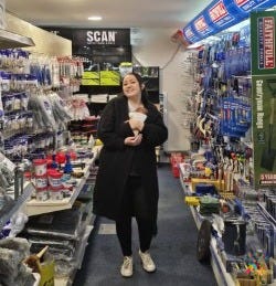 A woman in a long black coat and white trainers stand smiling whilst she holds a newborn baby in  the aisle of a well stocked builders' merchant. Shelves on either side display paint supplies, protective gear and hand tools. The store is brightly lit and neatly organised.