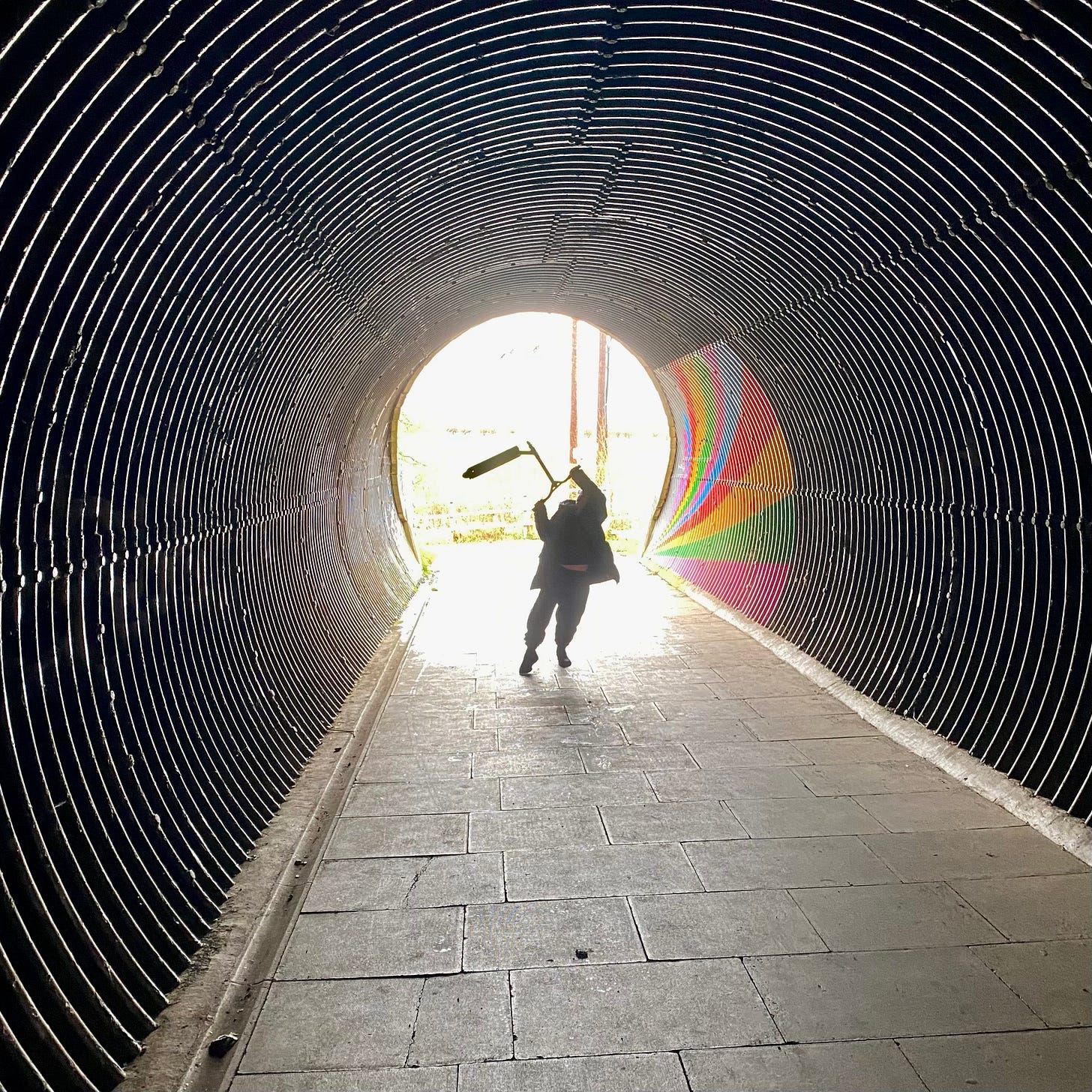 subway tunnel silhouette of boy waving scooter as if in triumph, rainbow pained on wall