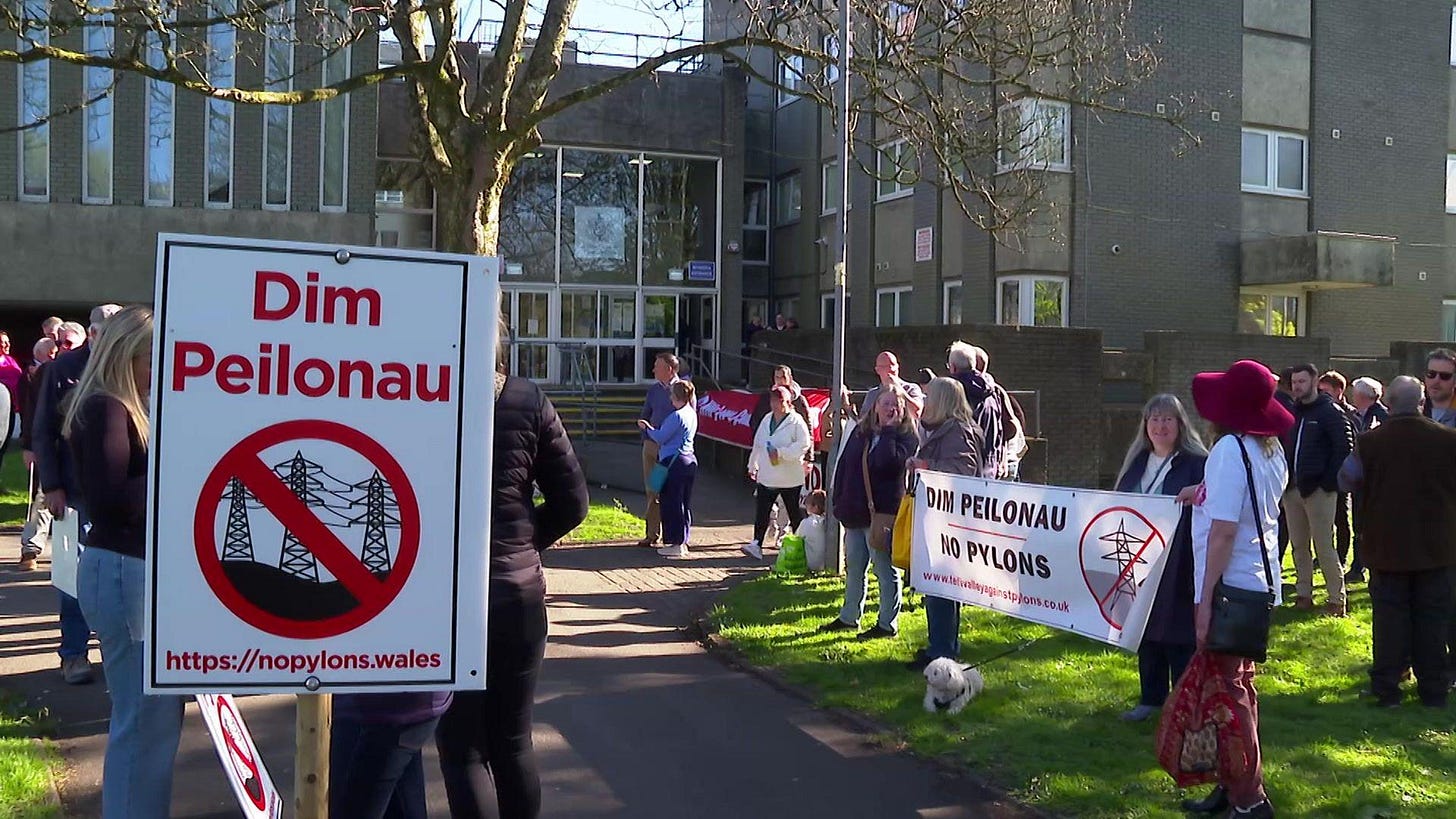 People are gathered outside a grey brick building holding banners with the words dim peilonau and no pylons.