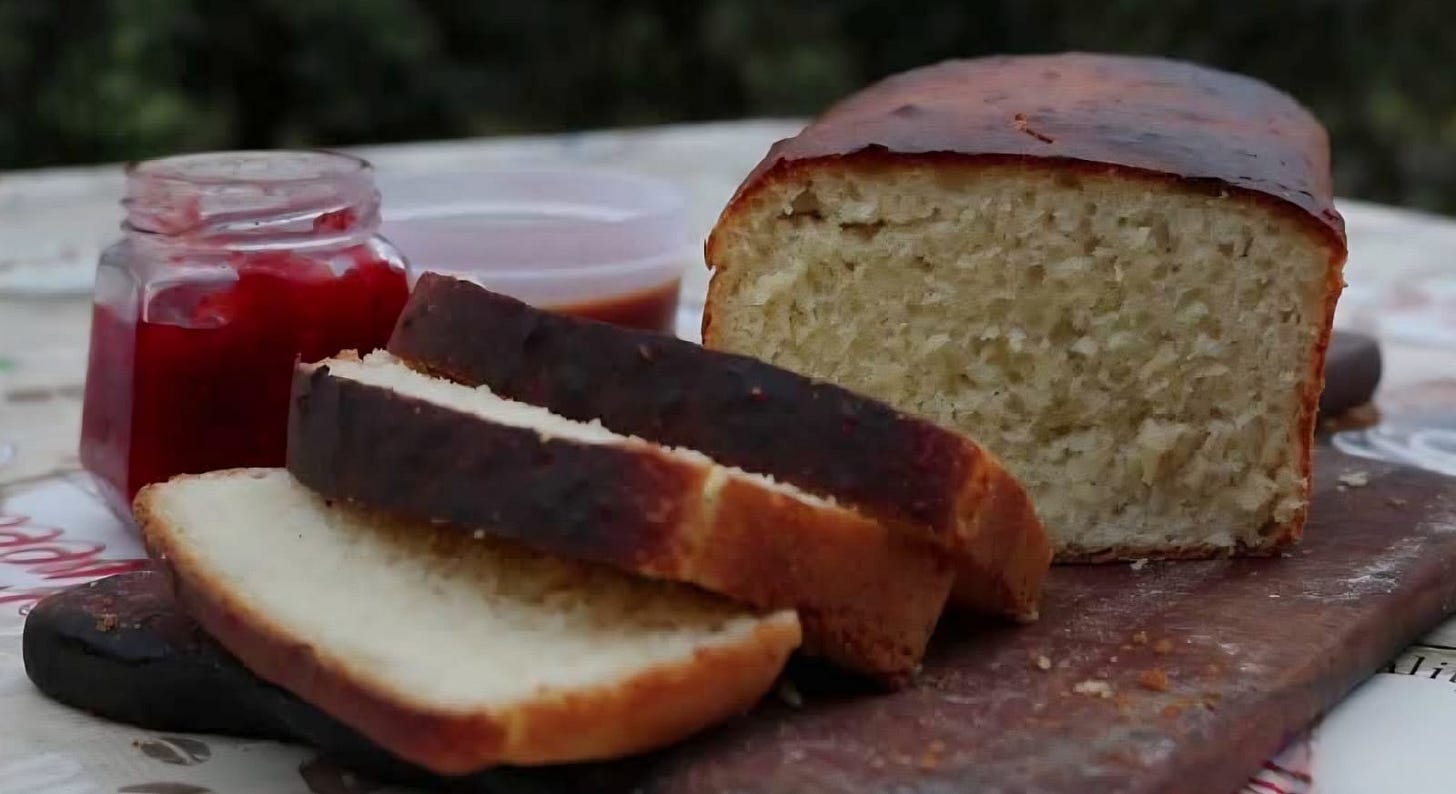 Pão caseiro de aipim fatiado sobre uma tábua de madeira com frasco de geleia ao lado