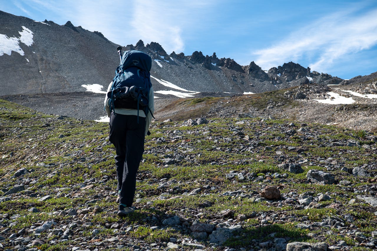 A hiker with a teal pack, seen from behind, walks up a gentle slope of green tundra dotted with grey rock toward a dark, jagged ridgeline. Patches of old snow cling to the slopes under a blue sky streaked with thin cloud.