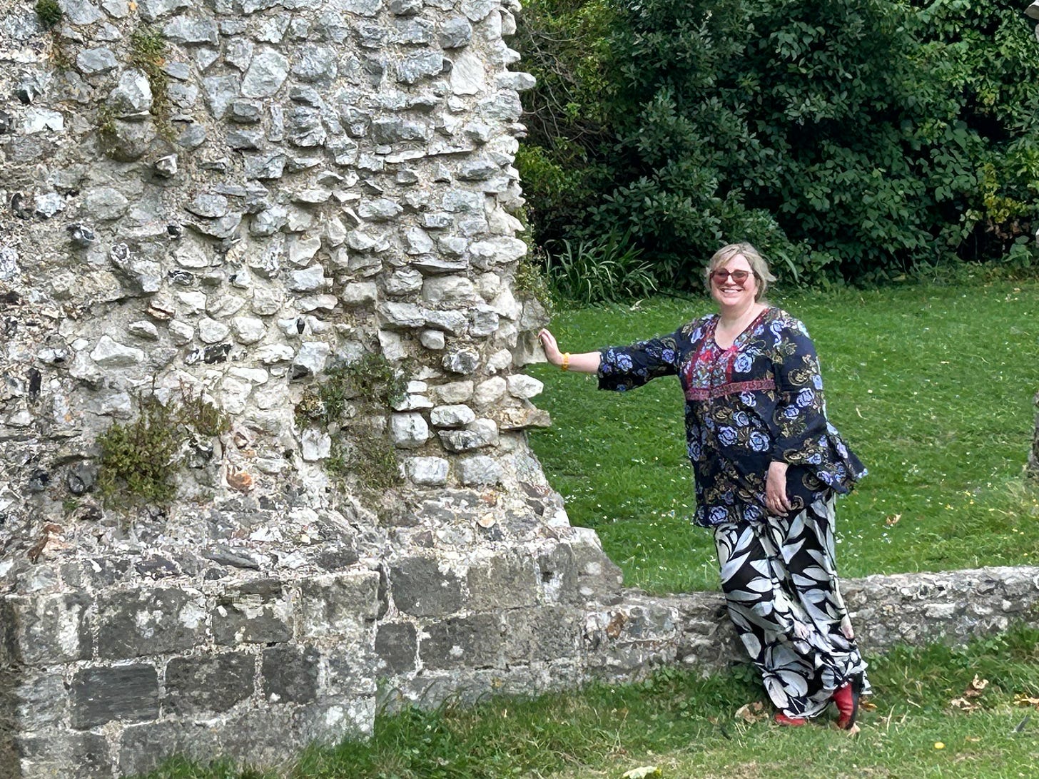 A white woman with blonde hair and colourful clothes stands with a hand against a wall of a ruined priory. A white woman with blonde hair and colourful clothes stands with a hand against a wall of a ruined priory.