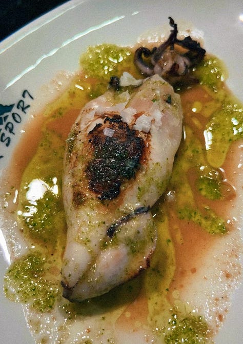 Crab stuffed squid, a spoon entering creamed uni in its shell, a hand holding a sesame crack with three different color pieces of smoked fish on top , a hand holding grilled foie on bread, a hand holding txistorra skewered onto a piece of bread, a fork entering a half eaten piece of cheesecake