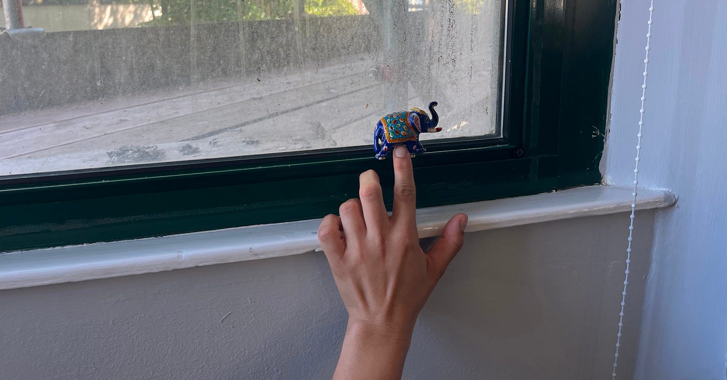 A hand reaches up to adjust a colorful elephant-shaped ornament on a dusty window frame. Natural light filters through the glass.