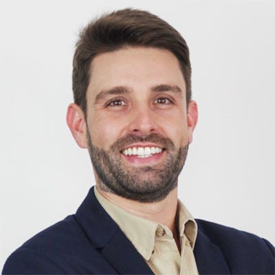Homem adulto, de pele clara, cabelos castanhos curtos e penteados para trás, e barba cheia e bem aparada. Veste uma camisa social bege sob um blazer azul-marinho. O fundo é liso, branco ou cinza-claro. Homem adulto, de pele clara, cabelos castanhos curtos e penteados para trás, e barba cheia e bem aparada. Veste uma camisa social bege sob um blazer azul-marinho. O fundo é liso, branco ou cinza-claro.