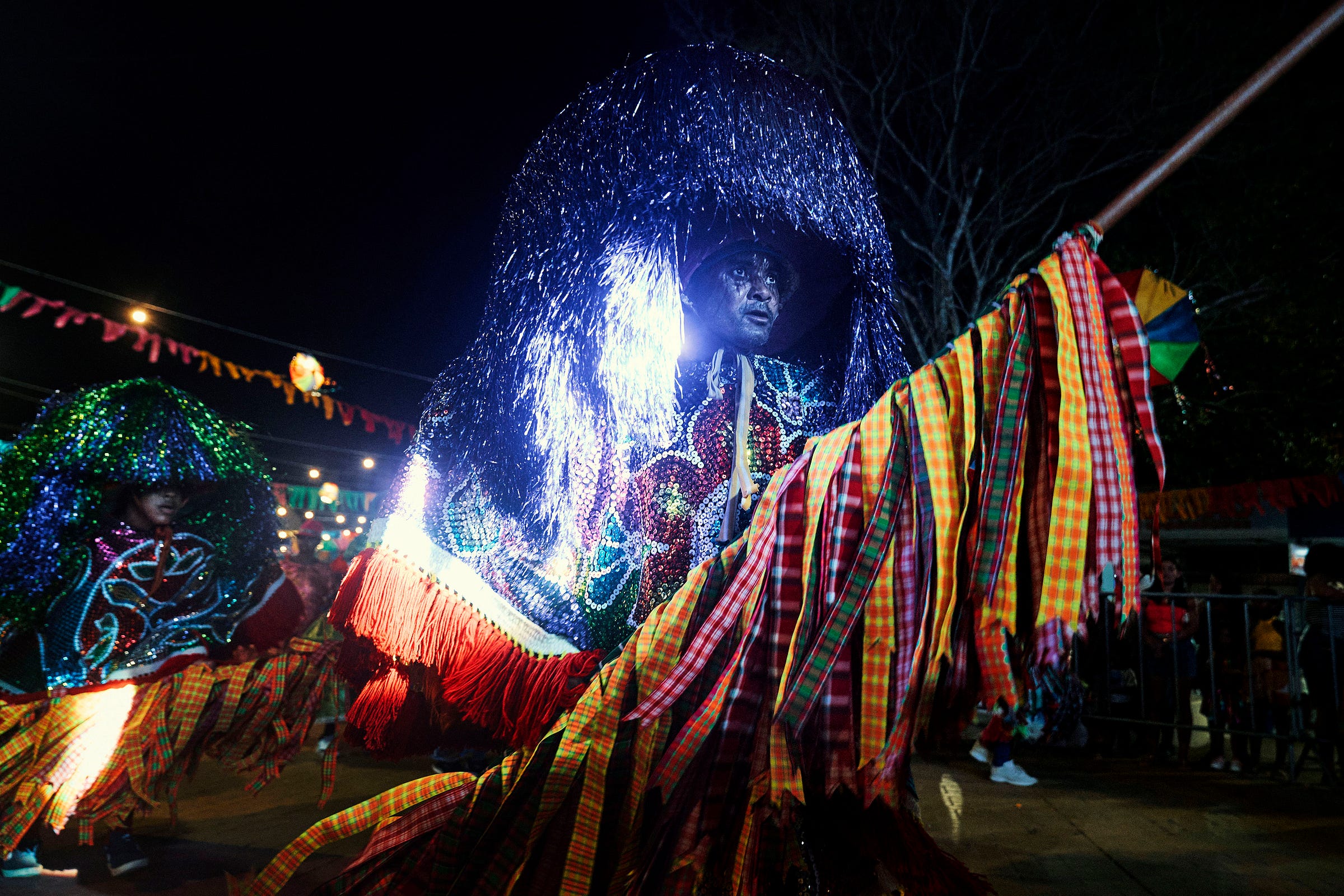 A person in a vibrant, sparkling costume and large headpiece participates in a nighttime parade, holding a colorful, fringed staff. Festive lights and decorations are visible in the background.