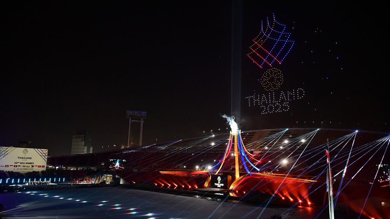 A nighttime view of the 33rd SEA Games opening ceremony at Rajamangala Stadium in Bangkok, featuring a drone display forming the Thailand 2025 logo above the arena, vibrant laser lights, and the illuminated central cauldron. A nighttime view of the 33rd SEA Games opening ceremony at Rajamangala Stadium in Bangkok, featuring a drone display forming the Thailand 2025 logo above the arena, vibrant laser lights, and the illuminated central cauldron.