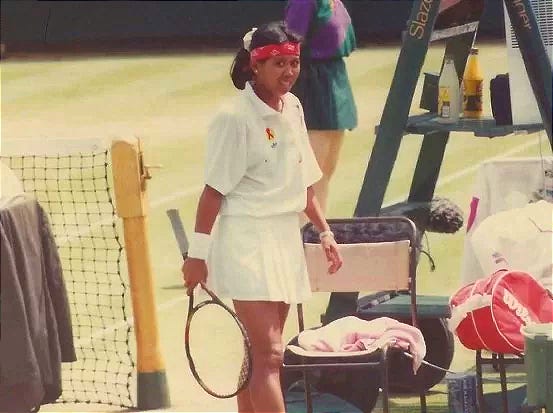 Indonesian tennis legend Yayuk Basuki on court during a match, dressed in a white tennis outfit with a red headband, holding a racket and smiling near the net. The photo captures a historic moment from her professional career, symbolizing Indonesia’s golden era in women’s tennis.