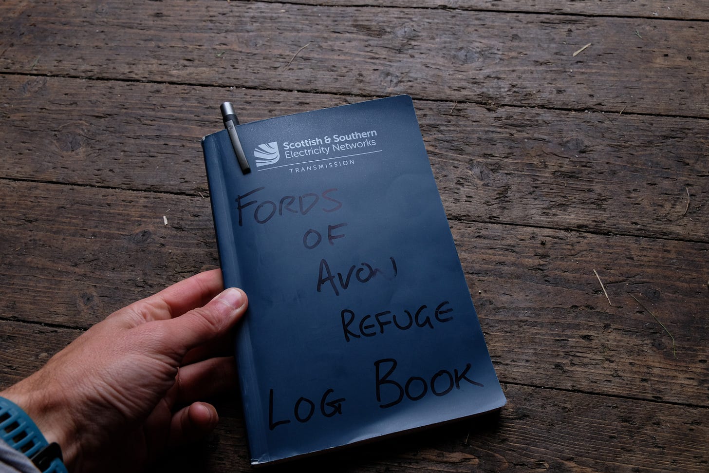 An open, well-used logbook rests on a wooden table inside a bothy. Its pages are filled with handwritten notes from visitors, recording years of shared experiences and memories.