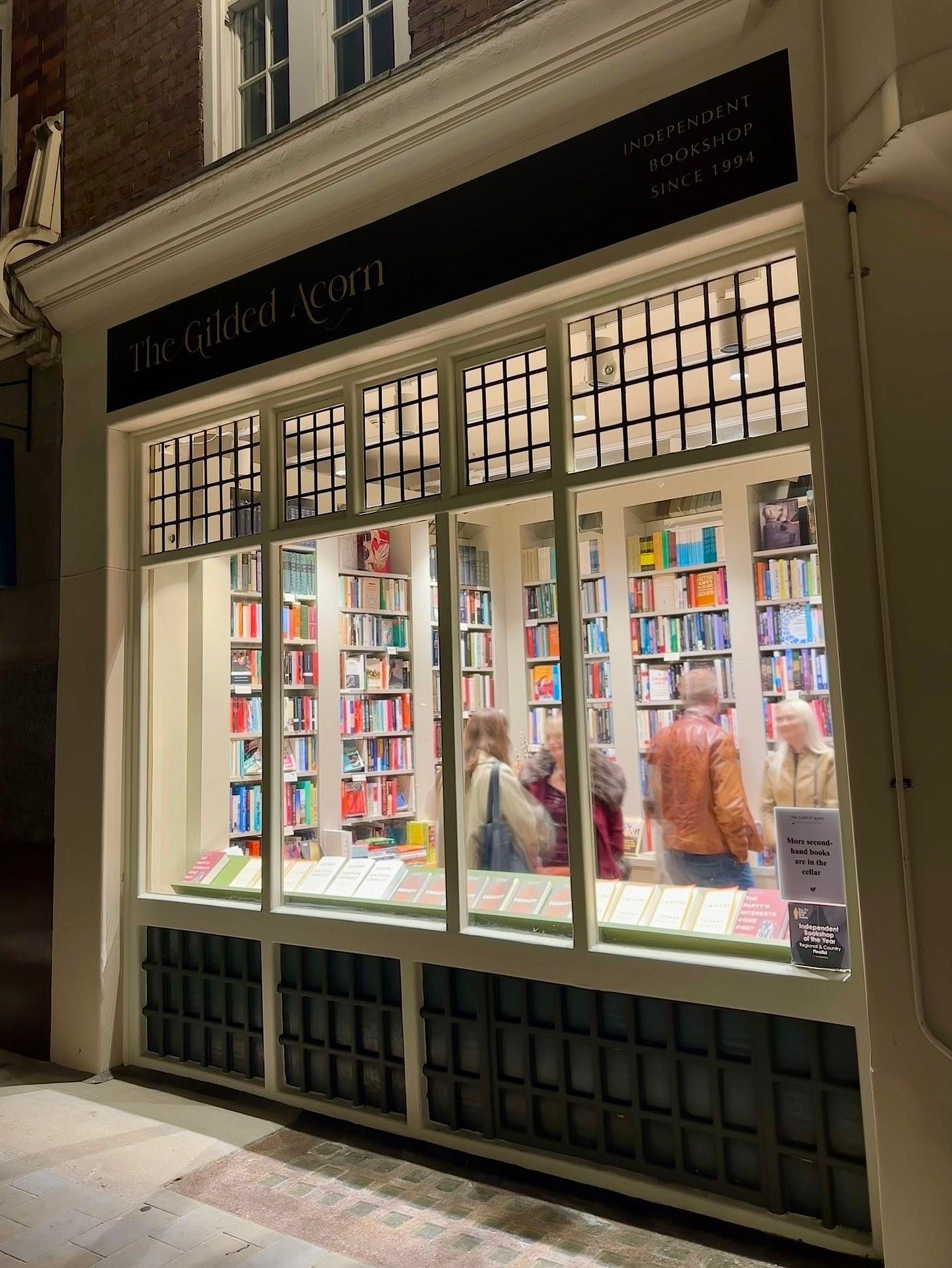 A long exposure shot taken from outside a tiny gem of an early twentieth century era bookshop in London called The Gilded Acorn. As dusk falls, the lights from inside the bookshop illuminate the courtyard outside and you can the blurred outline of a a few people inside. A long exposure shot taken from outside a tiny gem of an early twentieth century era bookshop in London called The Gilded Acorn. As dusk falls, the lights from inside the bookshop illuminate the courtyard outside and you can the blurred outline of a a few people inside.