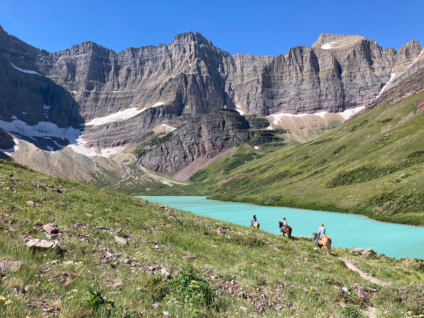 A jagged wall of mountain rock sits at the head of bright blue green lake while three horses with riders follow a trail. A jagged wall of mountain rock sits at the head of bright blue green lake while three horses with riders follow a trail.