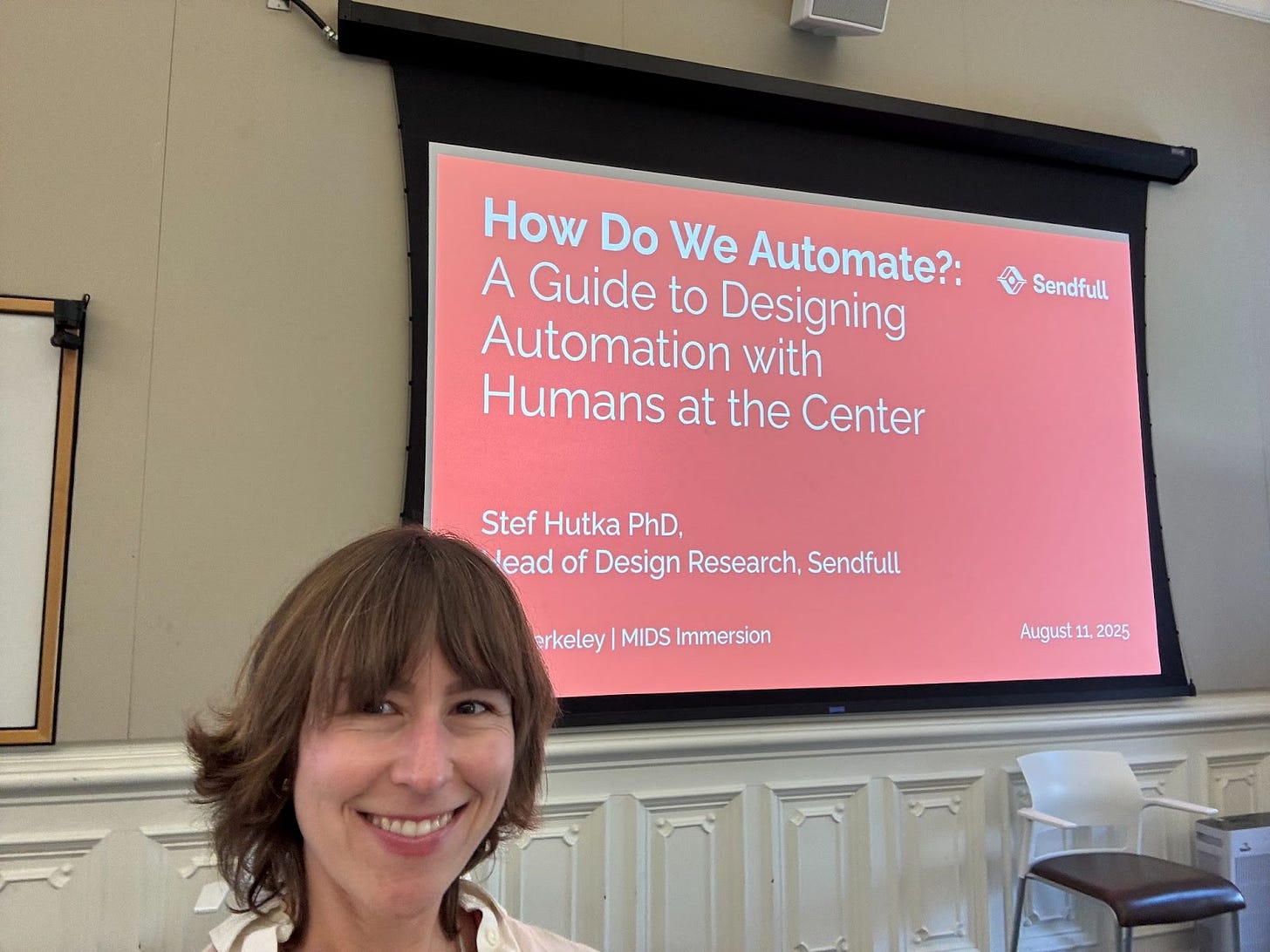 A woman smiling in a classroom with a presentation slide projected behind her. The slide reads: “How Do We Automate?: A Guide to Designing Automation with Humans at the Center” by Stef Hutka, PhD, Head of Design Research, Sendfull. The event is labeled “Berkeley | MIDS Immersion” with the date August 11, 2025.