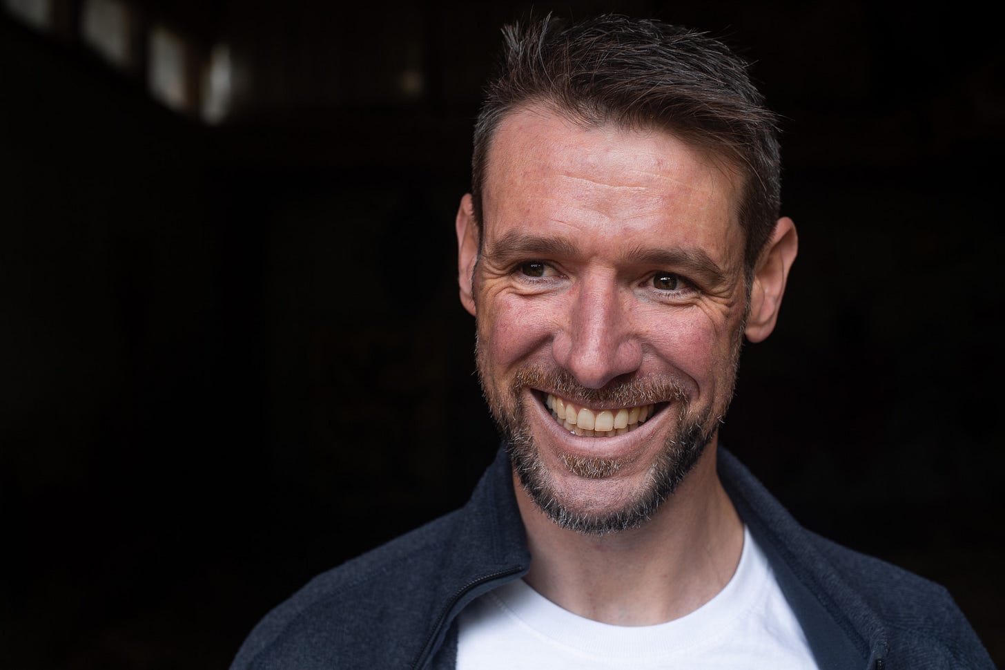 Adrian Hirt mit kurzem dunklem Haar und Bart steht vor einem dunklen Hintergrund. Sein Gesicht ist klar ausgeleuchtet, er trägt eine dunkle Jacke über einem hellen T-Shirt. Der Bildausschnitt zeigt ihn nah und frontal, während er leicht zur Seite blickt.