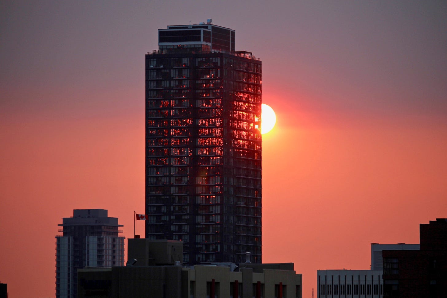 Bright sun peaking out from behind a skyscraper in Calgary creating a bright orange sky and lighting up the windows so they appear on fire. Also, a Canadian flag flying on a smaller building below.